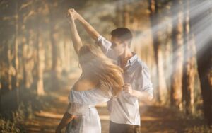 A young couple spinning and dancing together in a sun-drenched forest pathway.