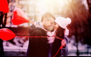 Smiling couple embracing outdoors in winter clothing next to red heart-shaped balloons.