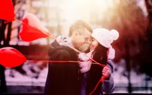 Smiling couple embracing outdoors in winter clothing next to red heart-shaped balloons.