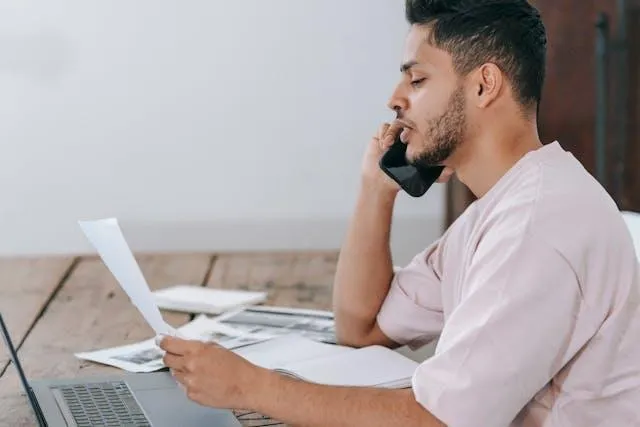 A man in a white shirt talking on the phone while looking at a piece of paper.