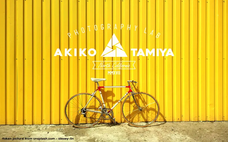 Yellow wall with a bicycle and a white vintage photography badge logo design reading Akiko Tamiya Photography Lab, North California.