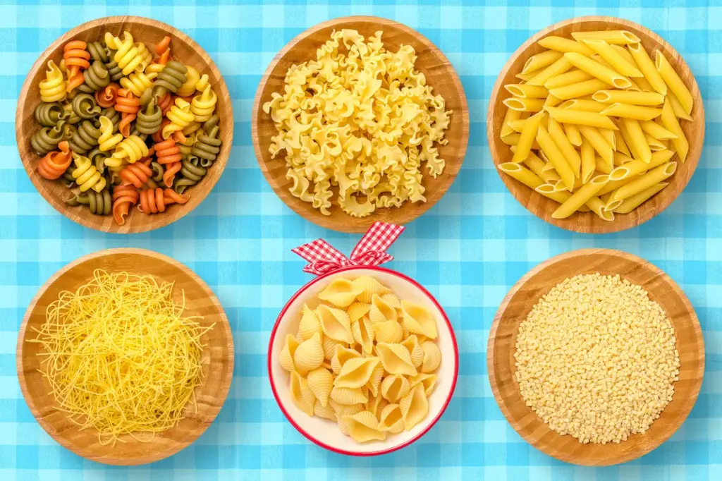 Seven bowls of dry pasta including rotini, mafaldine, penne, conchiglie, fideo, and acini di pepe on a blue checkered cloth