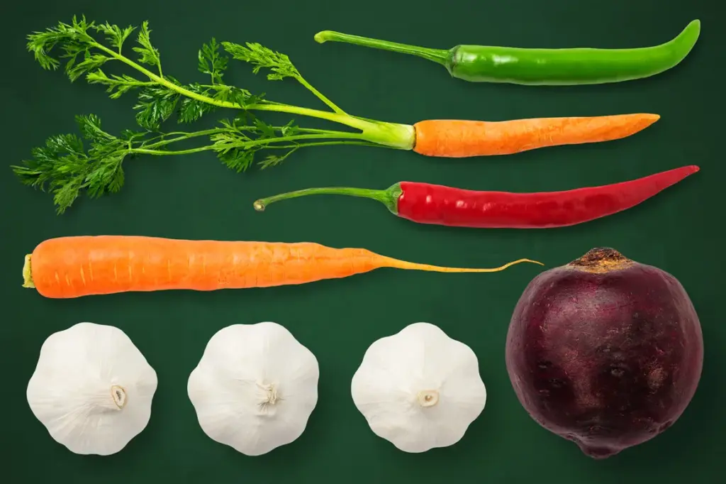 Green and red chili peppers, carrots, beetroot, and garlic bulbs arranged on a dark green background