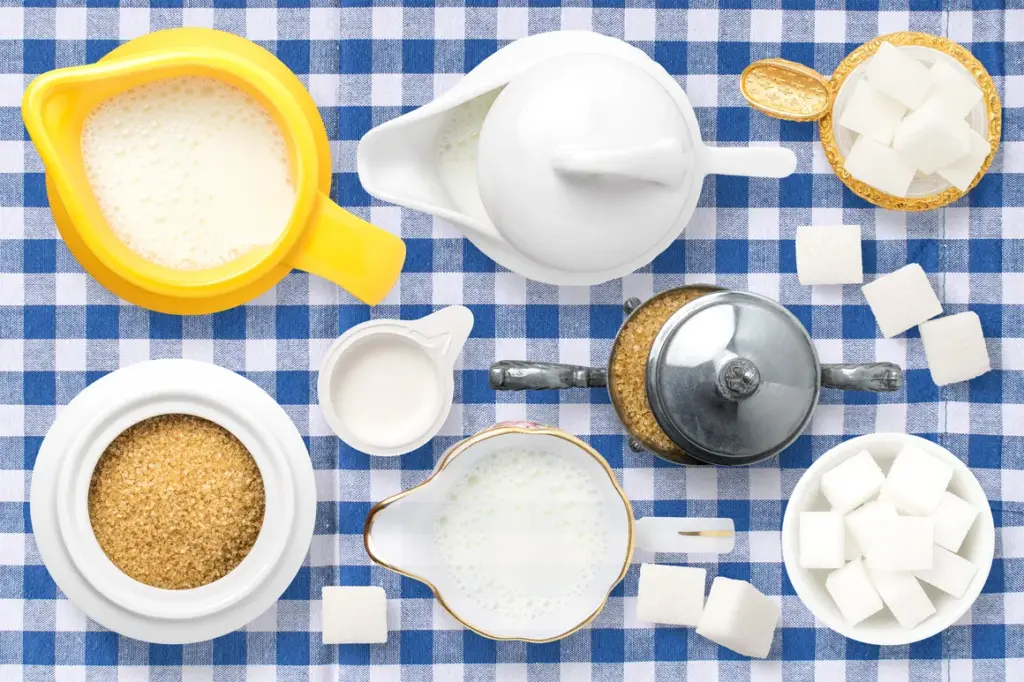 Pitchers and bowls of milk, brown sugar, and white sugar cubes arranged on a blue checkered tablecloth