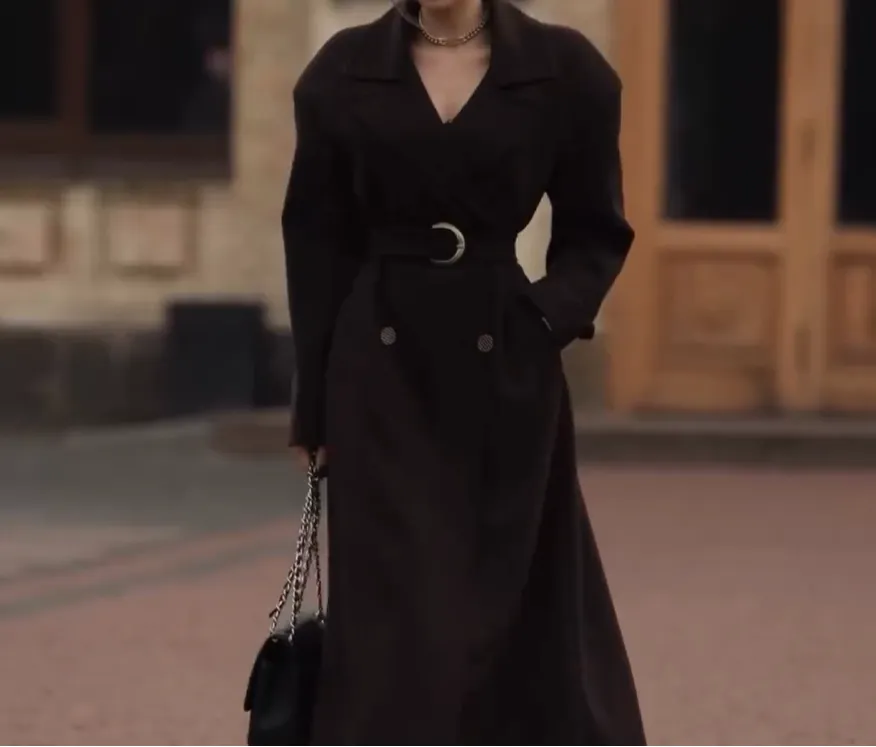 Woman wearing a long, dark belted trench coat walking outdoors.