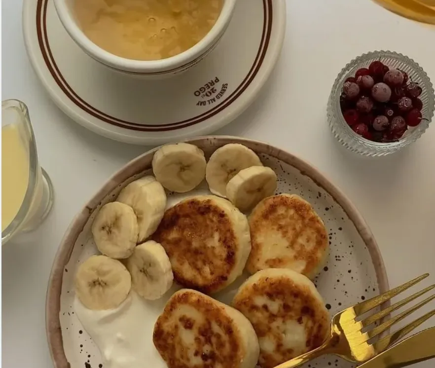 Overhead view of cheese pancakes (syrniki) with banana slices, sour cream, and a bowl of frozen berries.