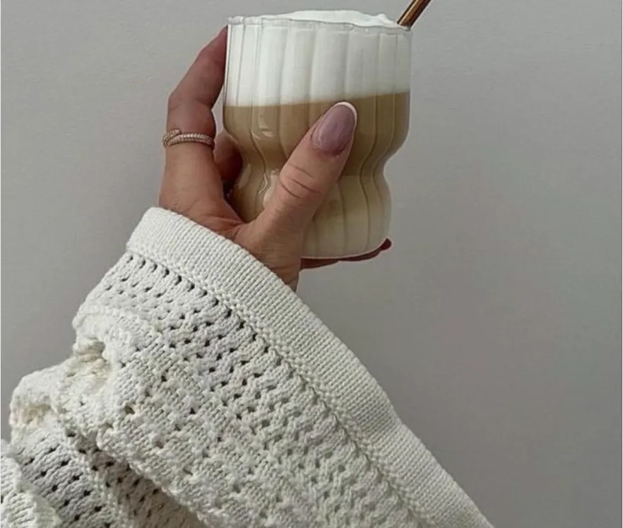 Hand in a white knitted sweater holding a fluted glass of layered coffee drink with foam.