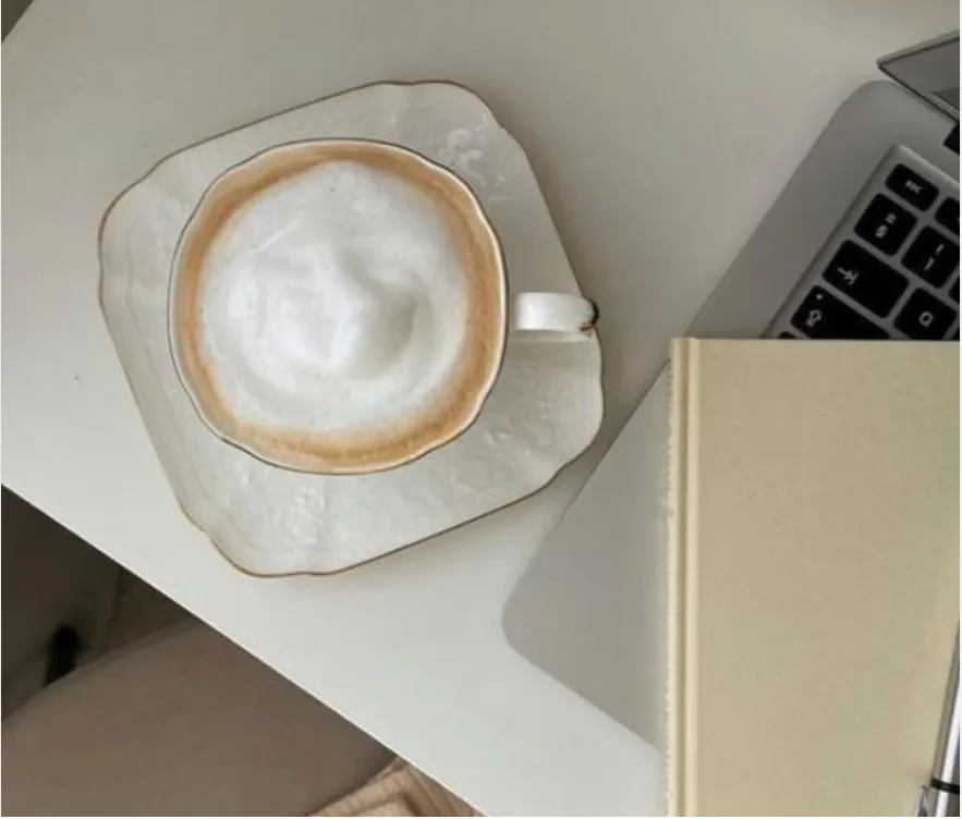 Overhead view of a cappuccino on a saucer next to a laptop and a blank notebook.