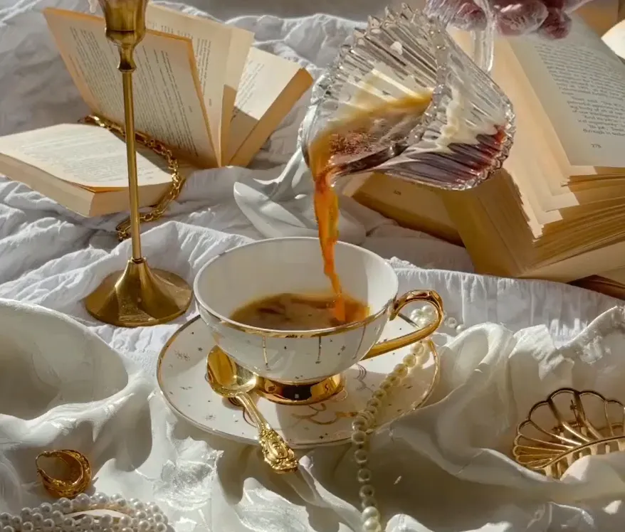 Close-up of coffee being poured into a gold-rimmed teacup surrounded by opened books and jewelry.