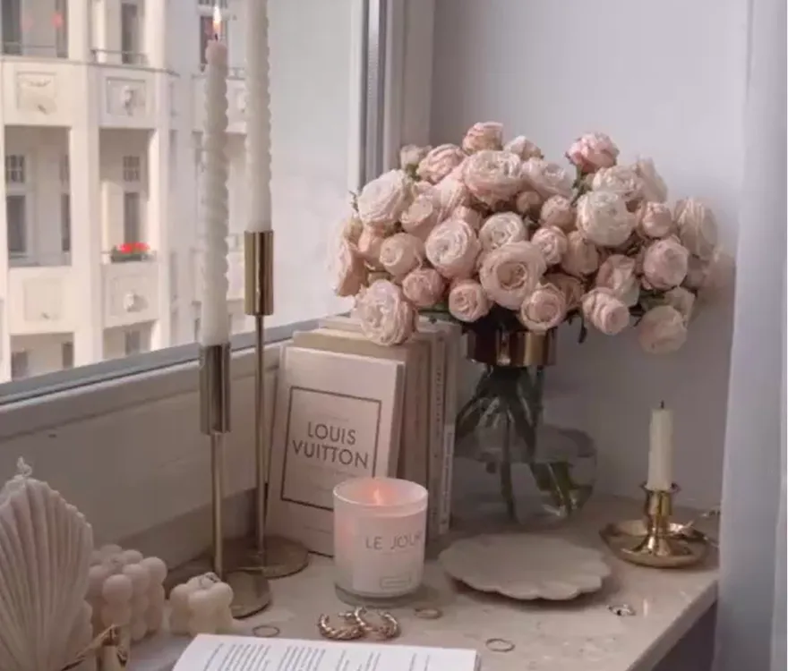 Window ledge decorated with pink roses in a vase, decorative candles, and fashion books.