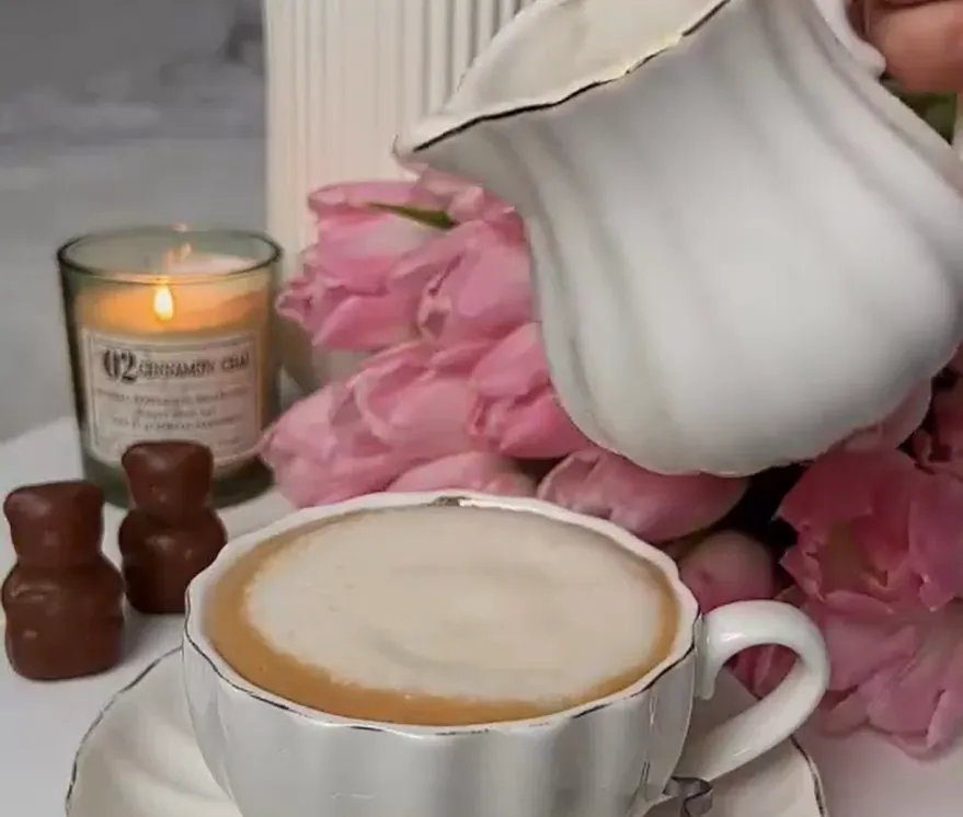 Hand pouring creamer from a white pitcher into a cup of coffee, with pink tulips and a lit candle nearby.