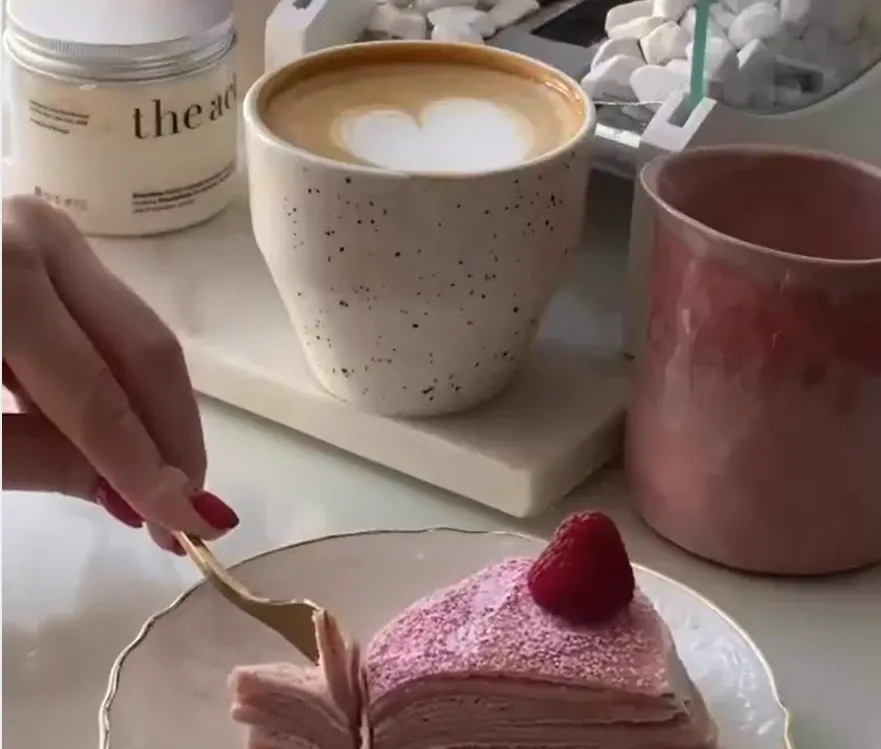 Hand cutting a slice of pink layered crepe cake topped with a raspberry next to a coffee with latte art.