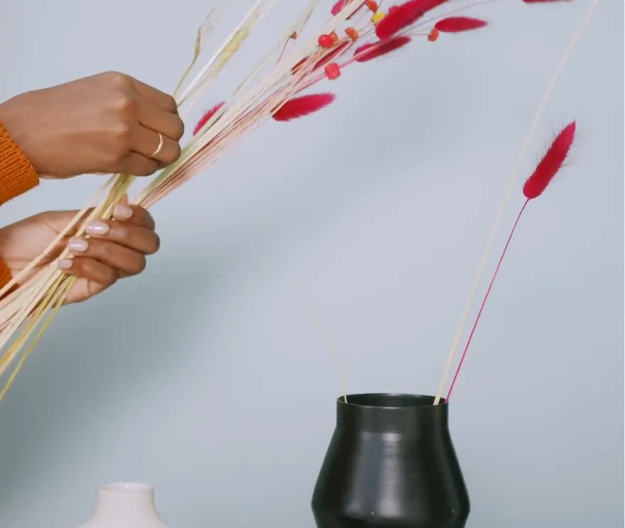 Hands arranging red and dried grasses in a dark vase.
