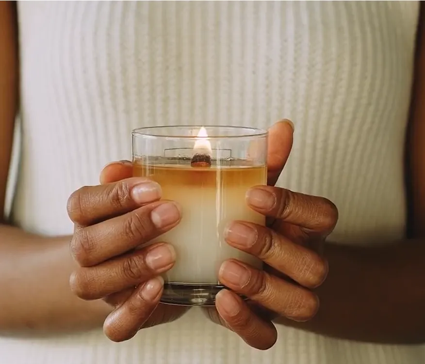 Hands holding a lit, layered candle in a clear glass jar.