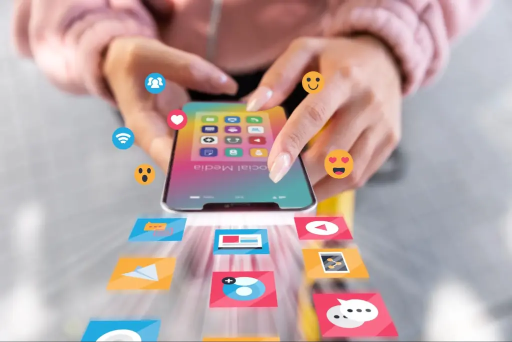 Close-up of a person's hands holding a smartphone, with colorful social media app icons and emojis digitally floating out of the screen.